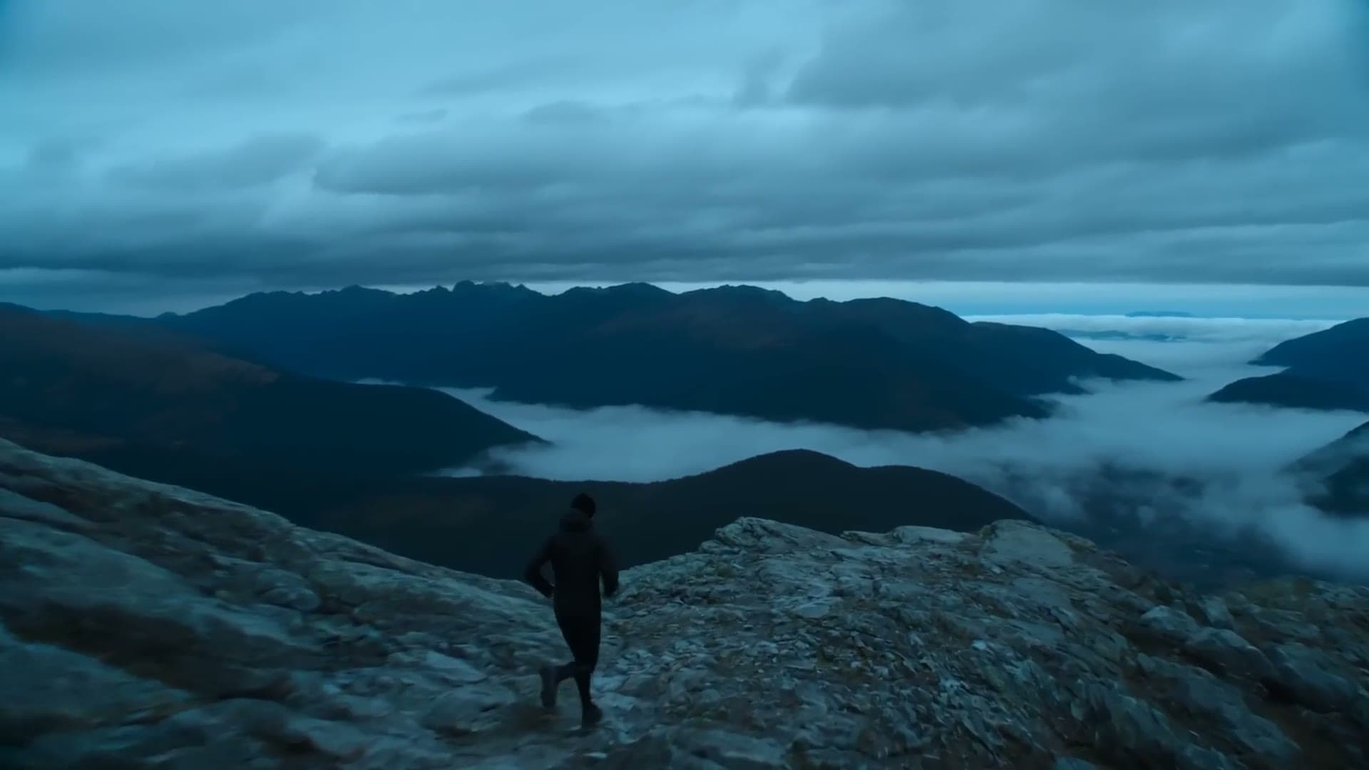 Trail runner moving along a rocky alpine ridge at blue hour, clouds pooling in the valleys below